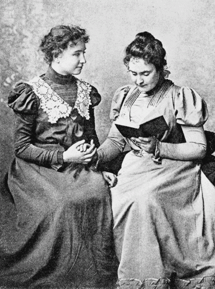 Helen Keller (to the left) and Anne Sullivan (to the right). Anne Sullivan reading a book to Helen who is .listening. by using her hand.